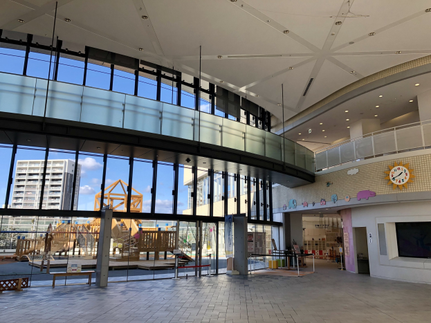 Innenansicht eines großen Gebäudes mit zahlreichen Fenstern, Deckenbeleuchtung, einer Wanduhr, einem Fernseher, einem Display, hölzernen Strukturen einschließlich eines Zauns, entfernten Gebäuden, Bäumen und einem bewölkten Himmel, die den Haupteingang zu einem neuen Kinderspielbereich markieren.