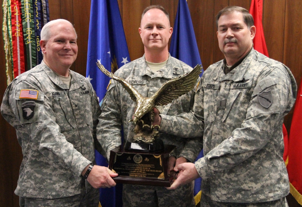 Drei Männer in Uniformen halten Auszeichnungen, mit Flaggen im Hintergrund und buntem Text in der linken oberen Ecke.
