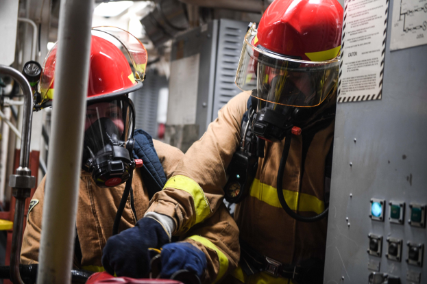 Zwei Feuerwehrleute in Schutzausröstung arbeiten an einem Feuerwehrauto, mit einer Tafel und Metallstäben im Hintergrund.