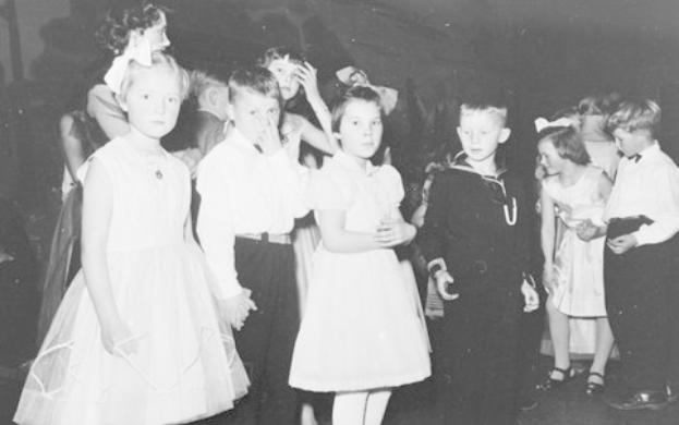 Schwarz-weißes Foto einer Gruppe von Kindern, die auf einer Tanzfläche stehen, lächeln und glücklich aussehen, mit einer Wand im Hintergrund.