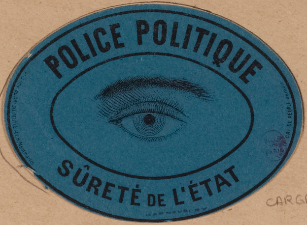 Ein brauner Karton mit einem blauen Aufkleber, auf dem "Polizei Politique Süreté de L'√Čtat" und ein Auge in der Mitte steht.