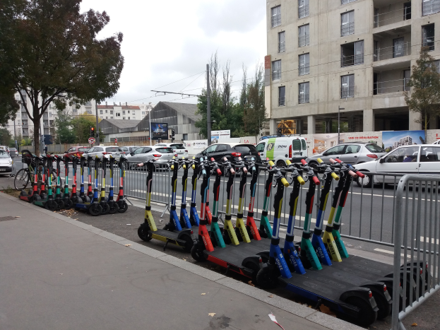 Eine Reihe von E-Scootern, die auf der Seite einer Straße mit einem Geländer auf der rechten Seite geparkt sind. Im Hintergrund gibt es Gebäude, Bäume, Pfosten, Verkehrszeichen und Schilder unter einem klaren blauen Himmel.