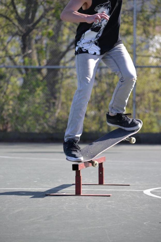 Eine Person in einem T-Shirt fährt Skateboard draußen mit einem Metallobjekt auf dem Boden, mit einem Netz, Metallstangen, Pflanzen und Bäumen im Hintergrund.