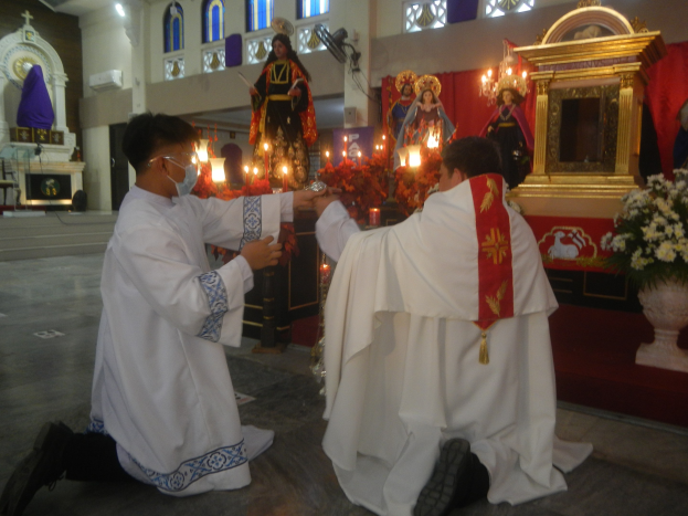 Zwei Priester knien vor einer Jesus-Statue, umgeben von brennenden Kerzen, mit Blumensträußen auf beiden Seiten des Altars und Menschen im Hintergrund.