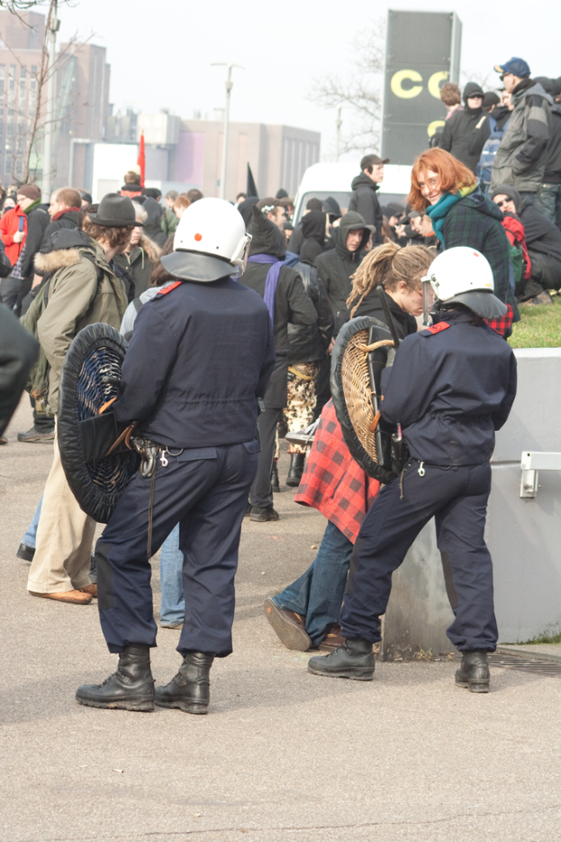 Eine Gruppe von Menschen auf einer Straße mit zwei Personen in der Mitte, die wie Polizisten aussehen, Gebäuden im Hintergrund und Boden unten.