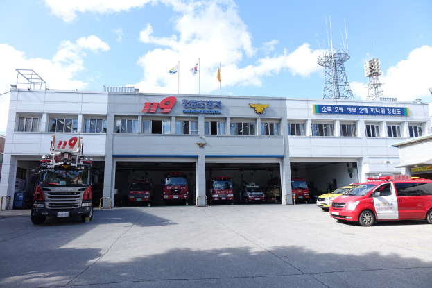Feuerwache mit mehreren Feuerwehren vor dem Gebäude, ein Gebäude mit Fenstern und Text, Flaggen an Stangen im Hintergrund, bewölkter Himmel und eine ferne Turm.