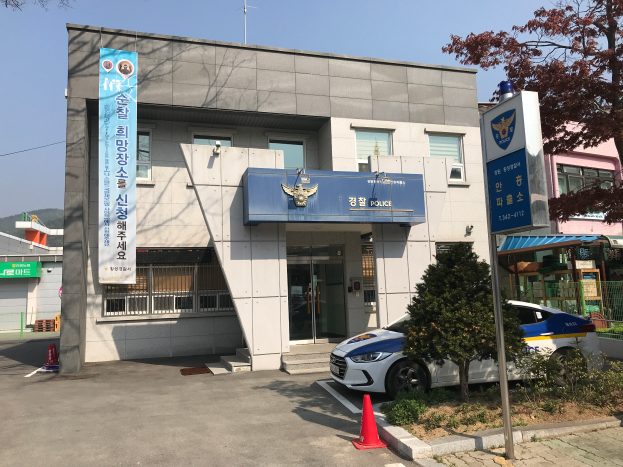 Ein Polizeiauto steht vor dem Seoul Police Headquarters, einem Gebäude mit Glasfenstern und -türen, umgeben von Bäumen, Pflanzen und Absperrbaken, mit einer Fahne mit Text auf dem Gebäude und Hügeln und einem klaren blauen Himmel im Hintergrund.