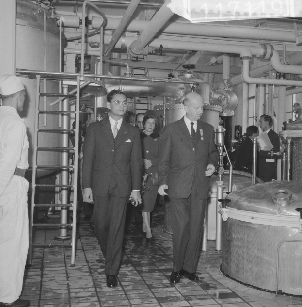 Eine Gruppe von Männern in Anzügen und Krawatten geht durch eine schwarz-weiße Fabrik, mit einer Leiter auf der linken Seite und Maschinen im Hintergrund.