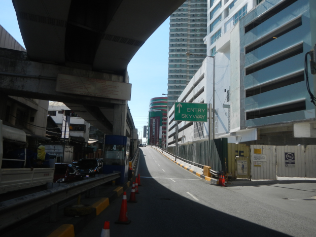 Eine Stadtstraße mit Verkehrskegeln am Straßenrand, Fahrzeuge auf der Straße, eine Brücke auf der linken Seite, Gebäude mit Namensschildern auf beiden Seiten, Laternenpfähle und ein Himmel im Hintergrund.