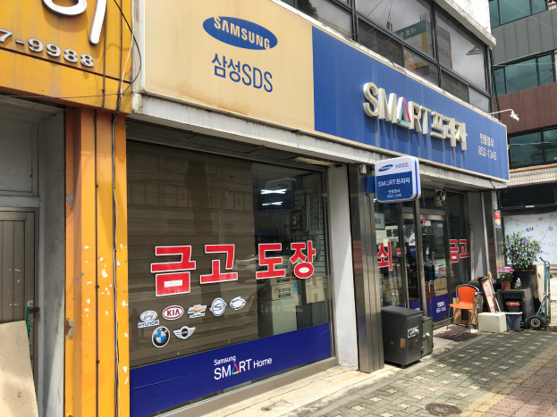 Ein Gebäude mit einer "Samsung Smart Home"-Aufschrift an der Seite, umgeben von anderen glaswandigen Gebäuden mit Namensschildern, Mülltonnen, Stühlen, Pflanzen und anderen Gegenständen, mit dem Himmel im Hintergrund.