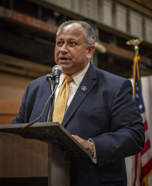 Ein Mann in Anzug und Krawatte steht an einem Podium mit Mikrofon und spricht, mit einer Flagge rechts und einer Wand dahinter, wahrscheinlich zu New York Citys neuem Bürgermeister.