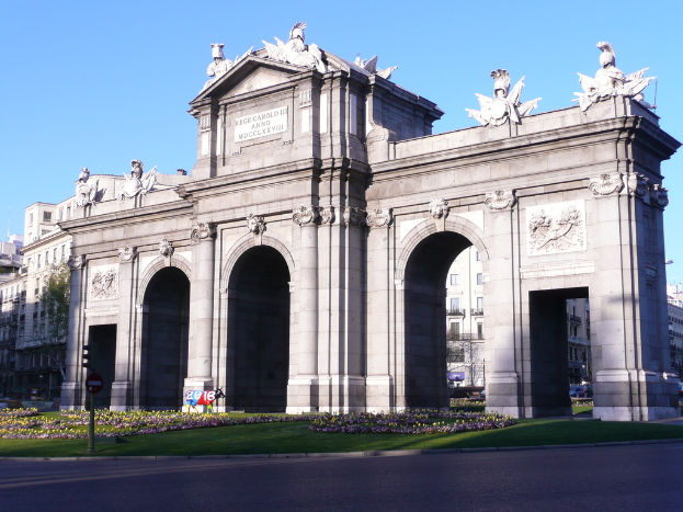 Gebäude und Skulpturen in der Mitte, mit einer Straße, Gras, Blumen, einem Pfahl und einer Tafel unten und Himmel oben.