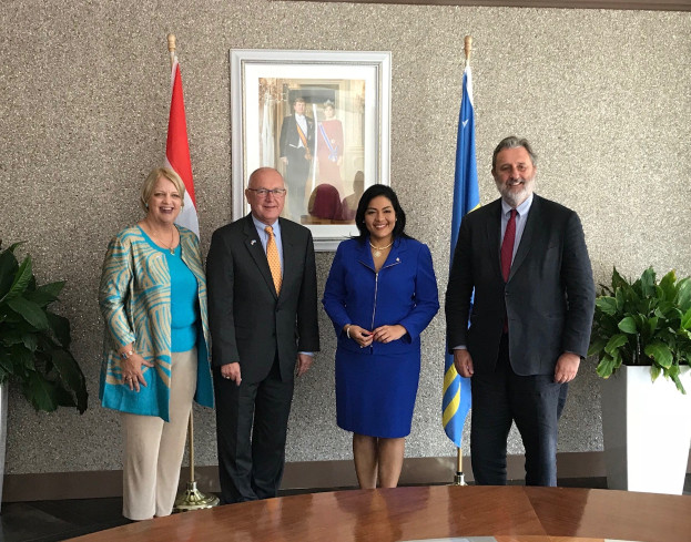 Gruppe von Menschen, die um einen Tisch mit Papieren stehen, umgeben von Fahnen, Topfpflanzen und einem gerahmten Foto an der Wand während einer Unterzeichnung.