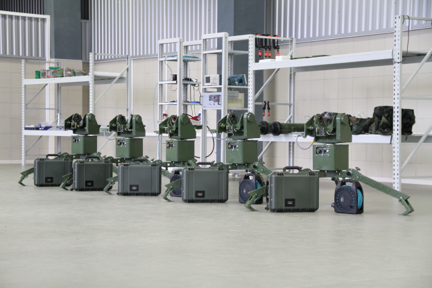 Eine Gruppe von Militärrobotern auf dem Boden, umgeben von Koffern und anderen Gegenständen auf Gestellen, mit einer Wand und einem Geländer im Hintergrund.