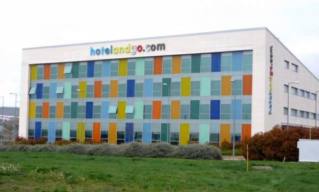 Großes Hotelgebäude mit vielen Fenstern, umgeben von Grünflächen, Fahrzeugen und einem bewölkten Himmel; Schilder lesen "hotel und go".