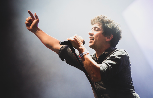 Ein Mann in schwarzem Hemd und Jeans singt in ein Mikrofon auf einer B├╝hne, mit geschlossenen Augen und einer jubelnden Menge im Hintergrund.