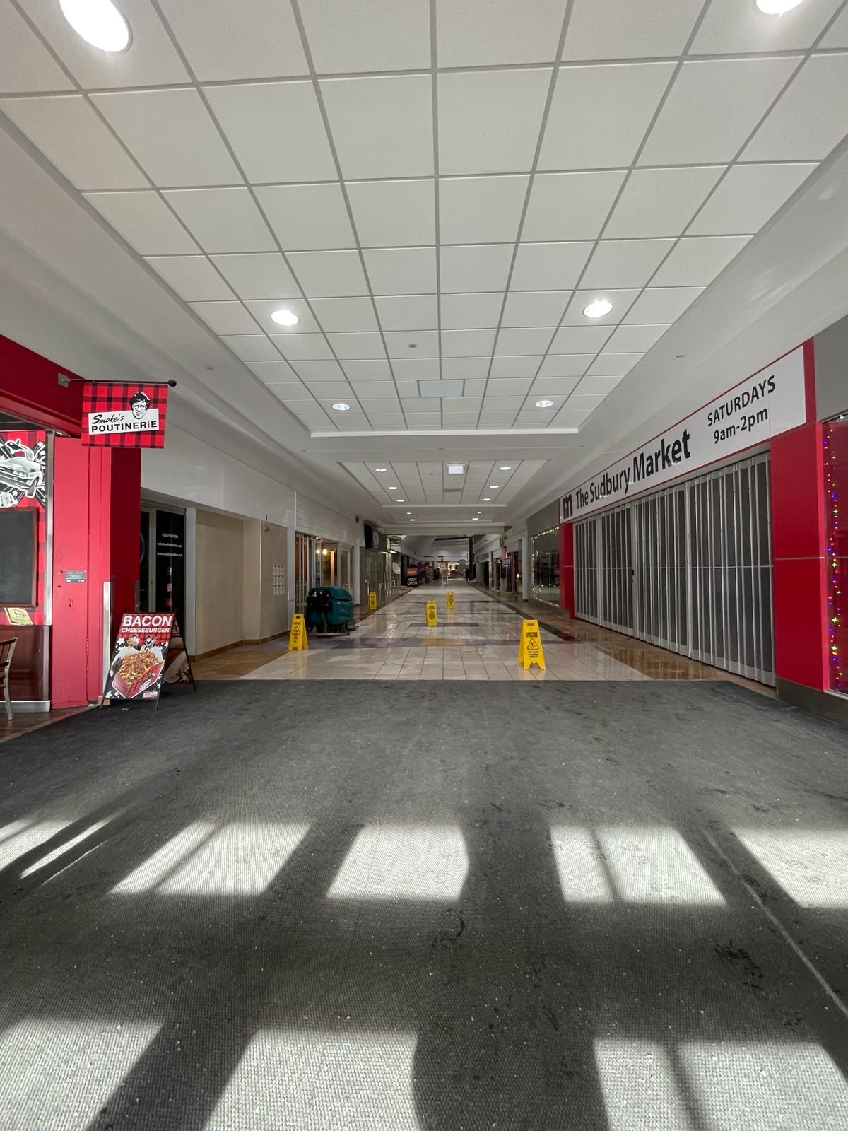 Ein hell erleuchteter Innenhof eines Einkaufszentrums mit Deckenbeleuchtung, einem Stuhl und einer Tafel mit der Aufschrift "Sydbury Market - Vorher und Nachher" links und verstreuten Gegenständen rechts und auf dem Boden.