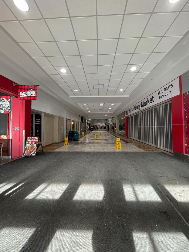 Ein hell erleuchteter Innenhof eines Einkaufszentrums mit Deckenbeleuchtung, einem Stuhl und einer Tafel mit der Aufschrift "Sydbury Market - Vorher und Nachher" links und verstreuten Gegenständen rechts und auf dem Boden.