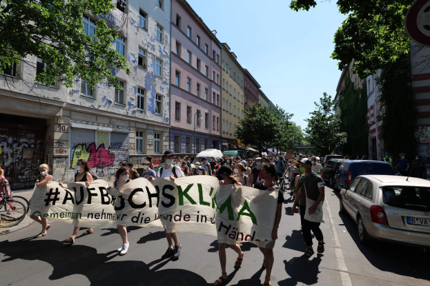 Eine Gruppe von Menschen marschiert auf einer Demonstration, hält ein Banner, mit parkenden Autos auf der rechten Seite und einem Radfahrer auf der linken Seite, vor einer Kulisse von Gebäuden, Bäumen und einem Schild in Berlin.