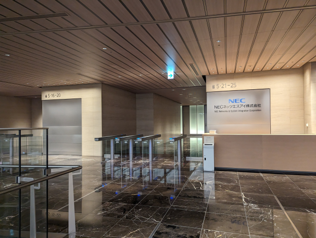 Eingangshalle eines Gebäudes mit Marmorboden, Glasgeländern, elektrischen Deckenleuchten, Namensschildern an den Wänden, einem Empfangstisch und einem Logo, das die Zugehörigkeit zur NEC Network & System Integration Corporation anzeigt.