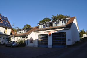Feuerwache mit Solarpaneelen auf dem Dach, umgeben von Gebäuden, Straßenlaternen, Fahrzeugen, Grünflächen und einem klaren blauen Himmel.