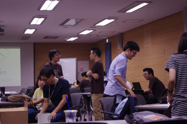 Ein Klassenzimmer mit Tischen, Stühlen, Laptops und Taschen; Menschen, die stehen und sprechen, ein Projektor, eine Holzwand und eine Tür im Hintergrund.