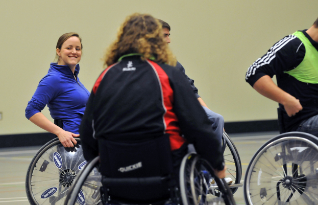 Eine Gruppe von Menschen sitzt in Rollstühlen auf dem Boden, mit einer Frau in einer blauen Jacke und einer Person in einem grünen Hemd, vor einer Wand im Hintergrund.