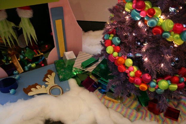 Eine Indoor-Szene mit einer Wand im Hintergrund, einem Leinen- und Wolltuch unten, einem geschmückten Weihnachtsbaum rechts, Geschenkboxen und Dekorationsgegenständen in der Mitte und einer Hütte mit Dekorationen oben links.