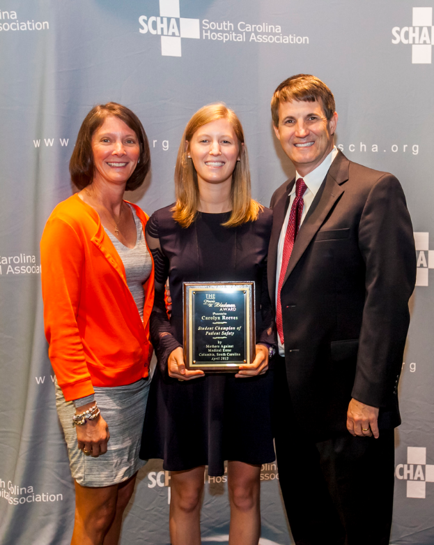 Drei Menschen, zwei Frauen und ein Mann, stehen zusammen; der Mann trägt einen schwarzen Anzug, eine Frau trägt ein orange-schwarzes Kleid, und die andere Frau, in einem schwarzen Kleid, hält einen Preis, mit einem grauen Hintergrund.