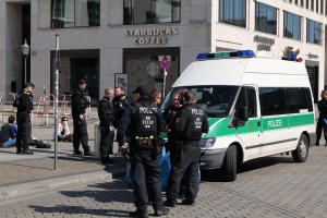 Eine Gruppe von Polizisten steht vor einem Starbucks-Café, rechts daneben parkt ein Van, links stehen und sitzen Menschen, im Hintergrund ein Gebäude mit Fenstern, ein Schild, ein Laternenpfahl und ein Zaun.