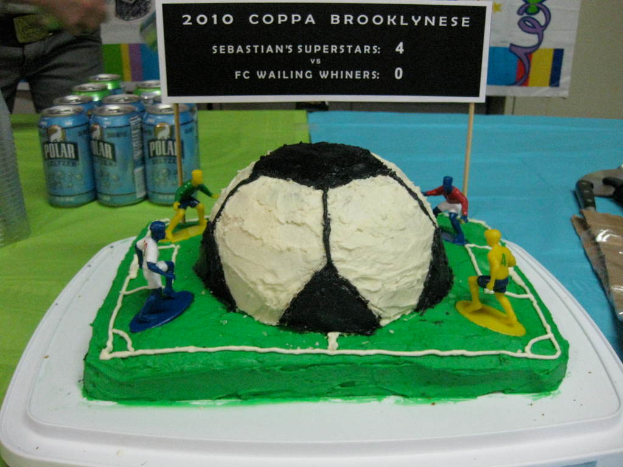 Ein Fußballstadion-Kuchen mit einem Fußball und vier Spielern, umgeben von Softdrink-Dosen, mit einem Banner im Hintergrund und einer Person oben links.