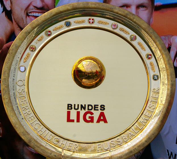 Goldener Schild mit "Bundesliga"-Text, gehalten von einem Mann, Hintergrund zeigt eine Fahne mit drei Personenbildern.