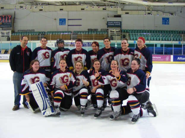 Eine Gruppe von Frauen-Eishockeyspielerinnen in Helmen und Schlittschuhen posiert für ein Foto auf dem Eis, eine hält einen Hockey-Schläger, mit Stühlen, Texttafeln, Geländern und Deckenlampen im Hintergrund.