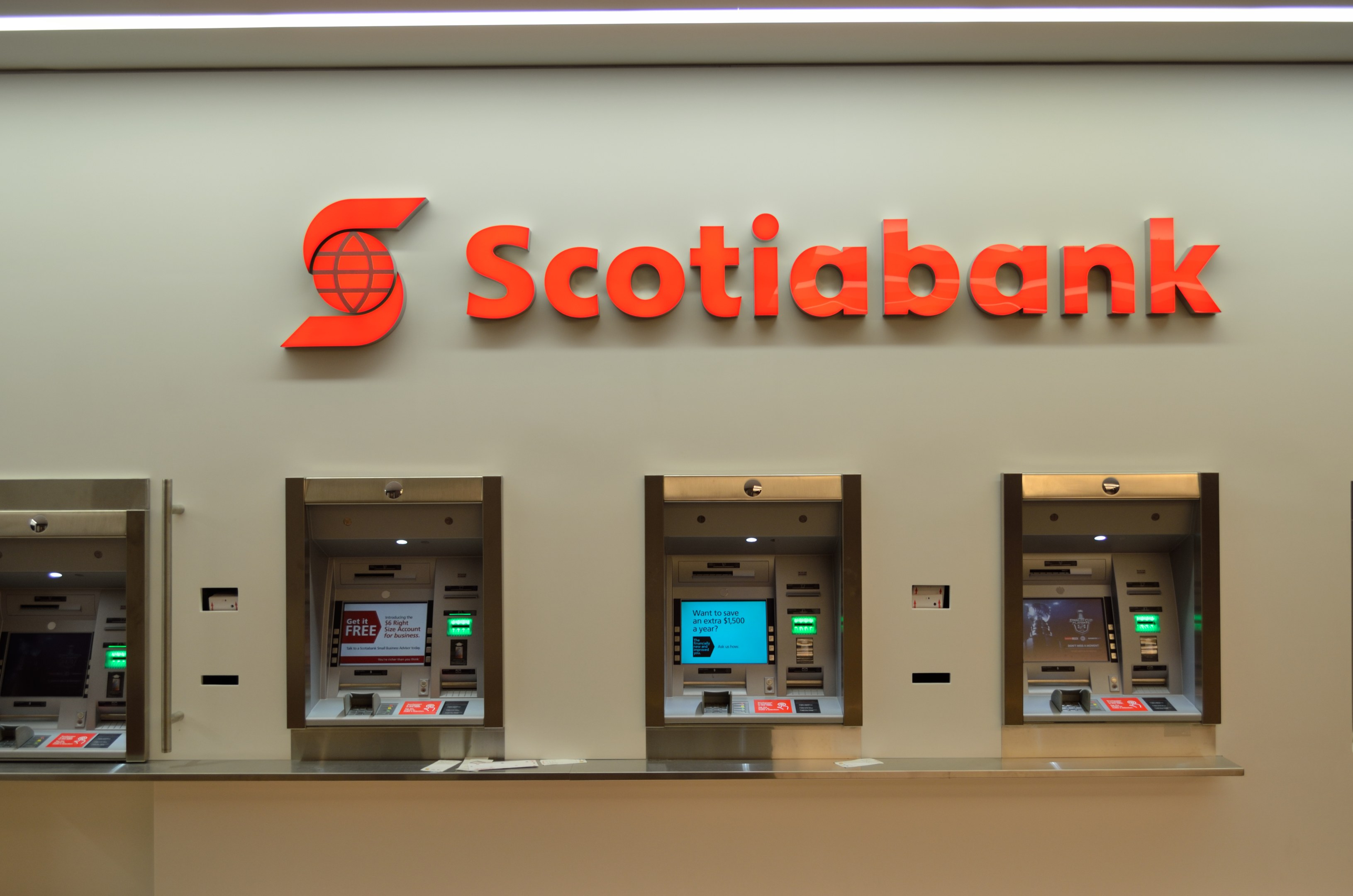 Ein modernes Scotiabank-Geldautomatengerät mit schwarzer und silberner Optik, einem großen Display und Bedientasten, vor einer hellgrauen Wand mit gerahmten Bildern und der Aufschrift "Scottish Bank" im Hintergrund.