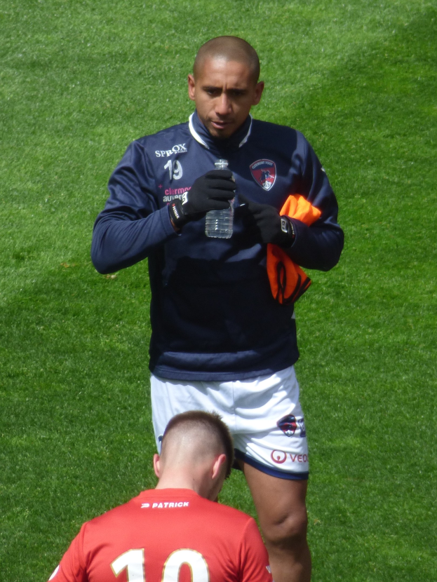 Ein professioneller Athlet in einem Sports Outfit steht auf einem saftig grünen Feld, in einer Hand hält er eine Flasche und in der anderen ein oranges Tuch.