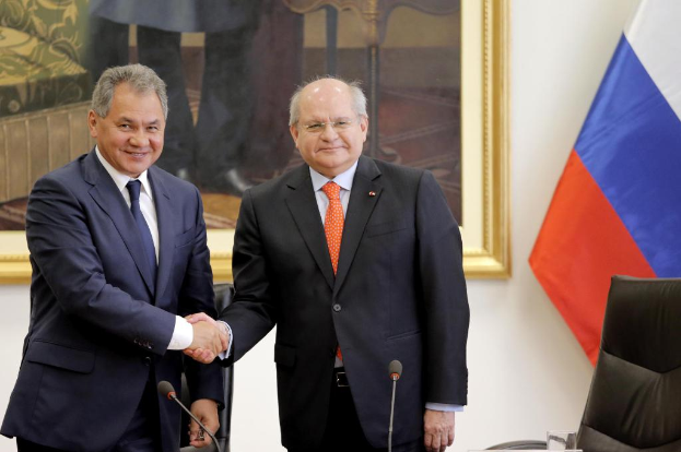 Zwei Männer im Blazer geben sich die Hand vor Mikrofonen, mit einem Stuhl, einer Fahne und einem Wandrahmen im Hintergrund.