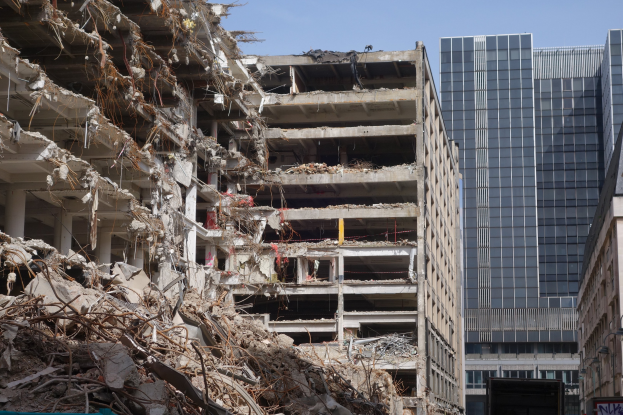 Ein große Abriss in einer Stadt umgeben von anderen Gebäuden, Fahrzeugen, Straßelementen, Bäumen und einem klaren Himmel.