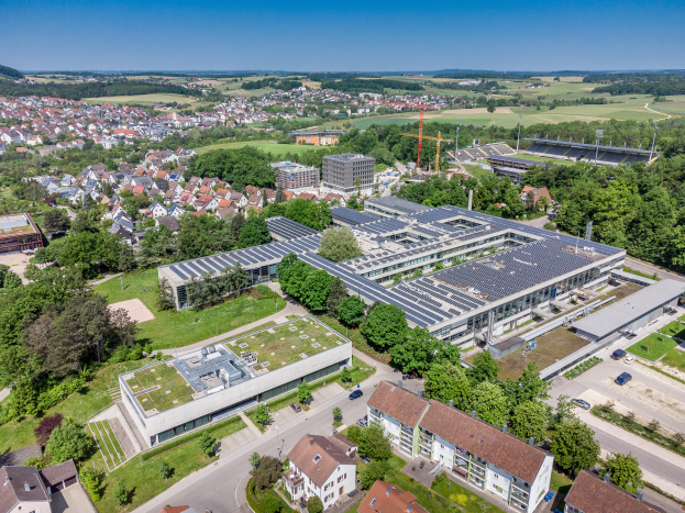 Luftaufnahme eines großen Gebäudes mit Solarpaneelen auf dem Dach, umgeben von Häusern, Bäumen und Fahrzeugen, mit Hügeln und einem klaren blauen Himmel im Hintergrund.