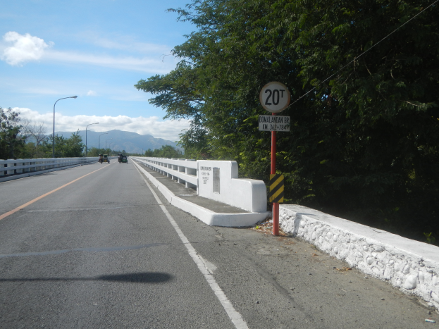 Eine Fahrbahn mit Fahrzeugen, ein Tempolimit-Schild, Geländer, Laternenpfähle, Bäume und ein bewölkter Himmel.