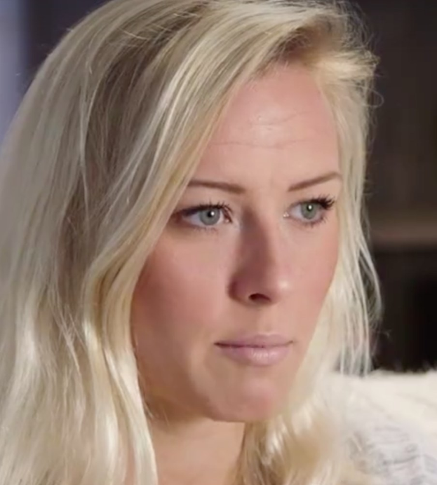 Blonde Frau mit blauen Augen sieht besorgt im Vordergrund vor einem unscharfen Hintergrund.