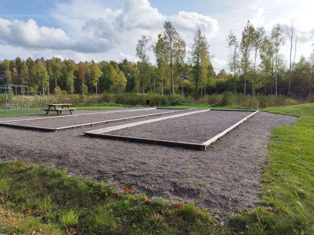 Eine grüne Wiese mit einem Picknicktisch, umgeben von Bäumen und einem klaren blauen Himmel, mit Spielplatzgeräten einschließlich Stangen und einer Bank auf der linken Seite.