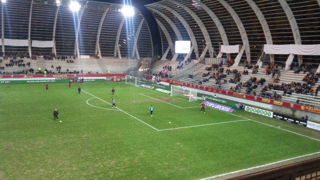 Ein Fußballspiel in einem großen Stadion mit Spielern auf dem Boden, einem Tor, eingezäunten Seitenlinien, Transparenten mit Text, einem Anzeigebildschirm, einem Dach mit Metallstangen und Deckenleuchten, unter einem bewölkten Himmel.