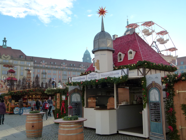 Ein geschäftiger Weihnachtsmarkt in Nürnberg, Deutschland, mit Menschen um dekorierte Stände, festliche Lichter und Schmuck, Gebäuden im Hintergrund, einem Riesenrad und einer Tafel auf der rechten Seite.