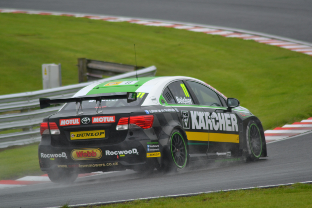 Ein Auto mit Text an der Seite auf einer regennassen Rennstrecke, umgeben von Gras, mit einem Geländer und einer hölzernen Konstruktion im Hintergrund.