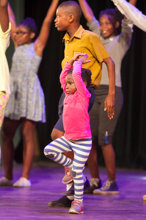 Eine Gruppe von Kindern auf einer Bühne mit einem Mädchen in der Mitte, das Schuhe trägt, vor einem dunklen Hintergrund.