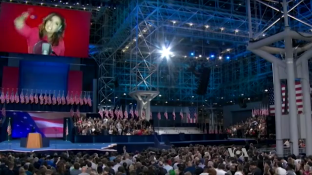 Eine große Menschenmenge sitzt vor einer Bühne mit einem Rednerpult, Mikrofon, Fahnen, einem Bildschirm mit dem Bild einer Frau und Lichtausrüstung, wahrscheinlich bei einer Präsidentschaftsdiskussion.