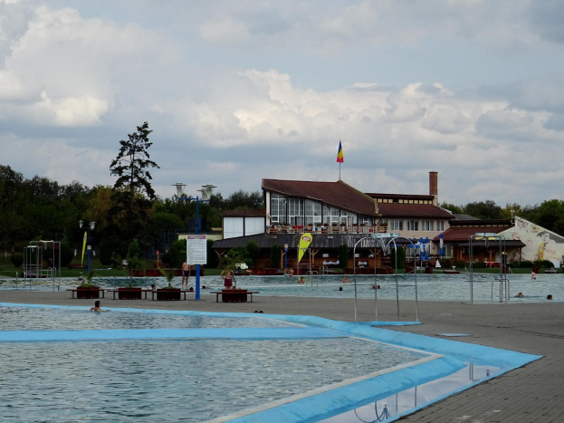 Ein großes Schwimmbecken voller Menschen, umgeben von Pfosten, Bänken, Topfpflanzen, einem Schild, einem Fahnenmast mit Flagge, einem Gebäude mit Fenstern, Straßenlaternen, einer Gruppe von Bäumen, unter einem bewölkten Himmel.