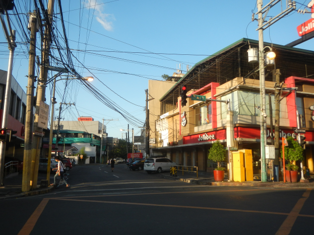 Eine Stadtstraßenszene mit Autos, Fußgängern, Strommasten, Laternen, Ampeln, Gebäuden mit Namensschildern, Topfpflanzen und einer bewölkten Himmel.