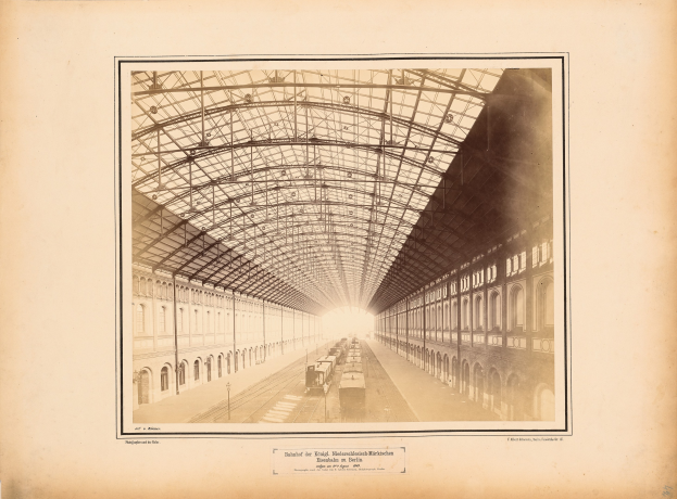 Schwarz-weiß-Foto eines Bahnhofs mit einem Zug auf den Schienen, umgeben von Gebäuden mit Fenstern und einem Dach, das von Metallpfosten gestützt wird; Text ist unten im Bild sichtbar.