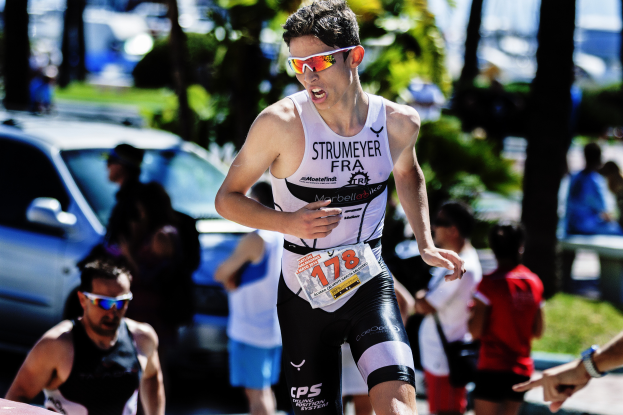Ein Mann in einem Triathlonanzug und Sonnenbrille läuft eine Straße entlang, umgeben von Menschen, Autos, Bäumen und Pflanzen, mit einem unscharfen Hintergrund, der Bewegung suggeriert.
